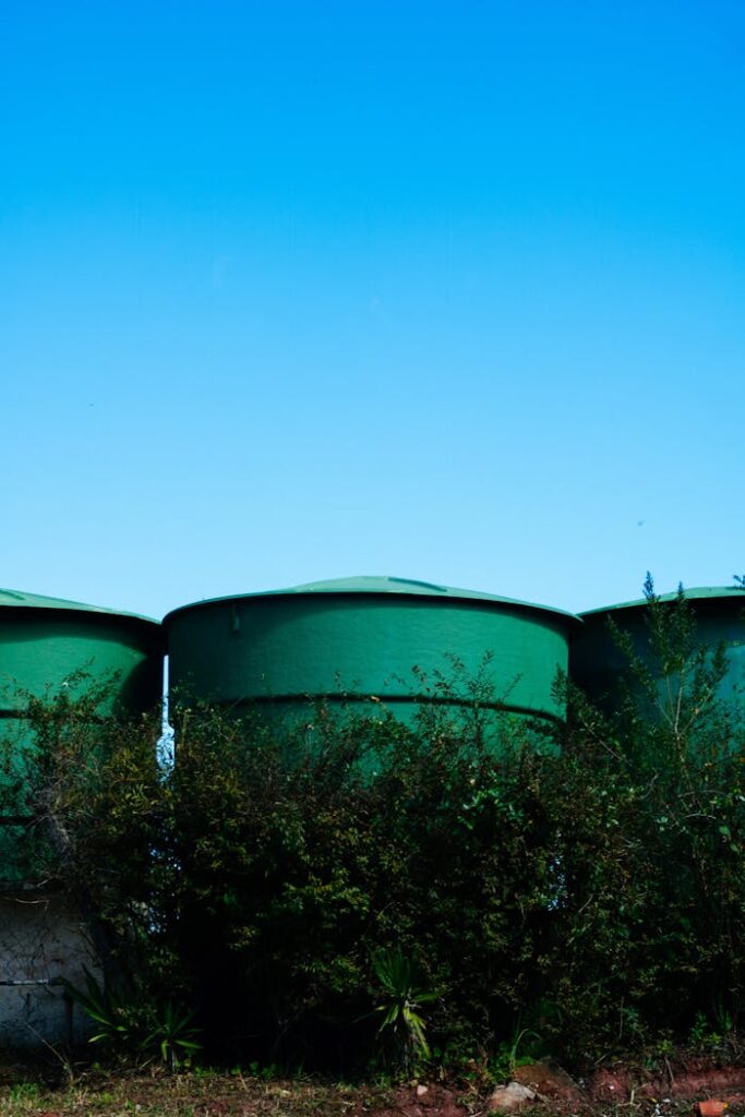 Green cylindrical water tanks in a rural area under a clear blue sky, surrounded by bushes.